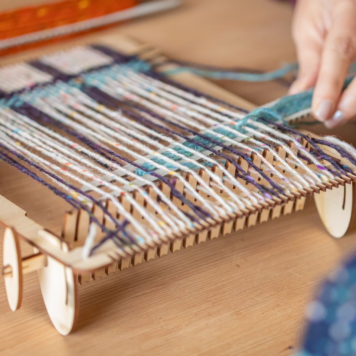 Hands weaving colourful yarn on a wooden Rocket Loom, with a Pattern Maker lifting the warp threads.